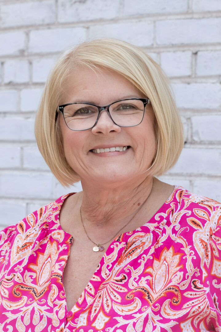 A woman with blonde hair wearing glasses and a pink shirt.